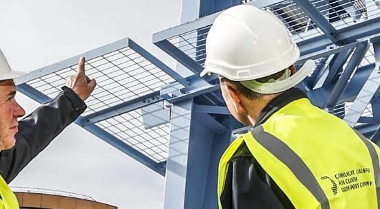 Two men in construction PPE wearing Dublin Port Company hi viz vests stand and point at a structure