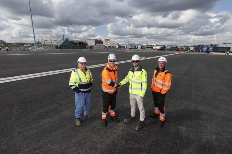 Four construction workers in high-visibility jackets and hard hats stand on a newly paved area at Dublin Port Terminal 4, with one pair shaking hands, and trailers and port infrastructure visible in the background under a partly cloudy sky.