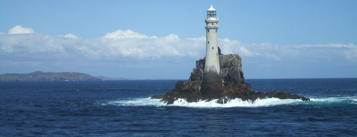 Fastnet Rock Lighthouse standing on a small rocky island surrounded by the ocean, with distant land visible on the horizon.