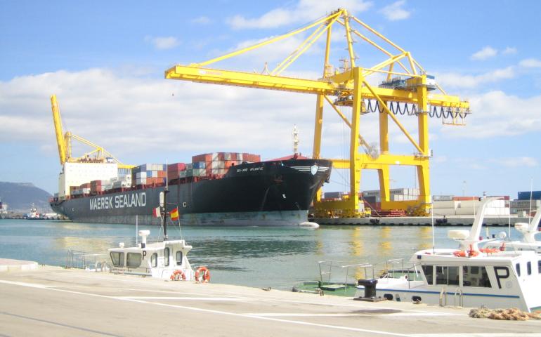Container ship docked at a port with yellow gantry cranes in the background and smaller boats moored in the foreground.