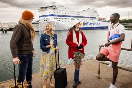Four individuals dressed in various holiday outfits stand before a Brittany Ferries vessel