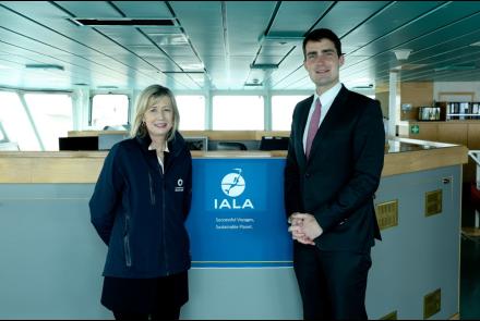 Two individuals standing in front of the IALA logo