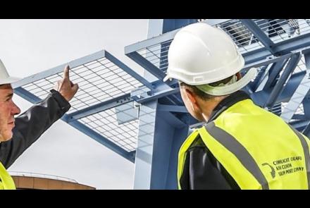 Two men in construction PPE wearing Dublin Port Company hi viz vests stand and point at a structure