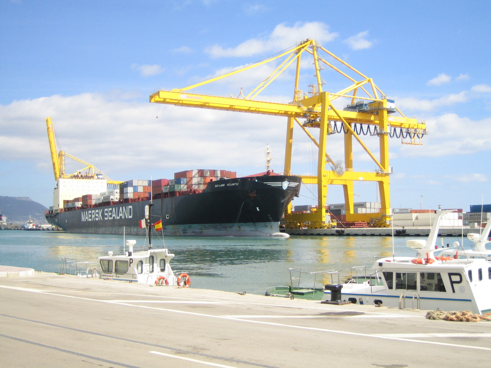 Container ship docked at a port with yellow gantry cranes in the background and smaller boats moored in the foreground.